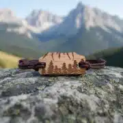 Holzarmband – Mittagszähne – 1 7-Gipfel-Holzarmband mit eingraviertem Motiv der Dents-du-Midi-Berge und Tannen, geflochtene braune Kordel, auf nassem Stein mit schneebedeckter Bergkette im Hintergrund