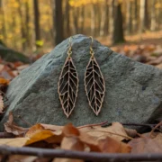 Boucles-oreilles-en-bois-ekinox-4 Boucles d'oreilles en bois Ekinox avec pendentifs feuilles en bois suisse sur crochets dorés, posées sur pierre grise avec feuilles d'automne et forêt en arrière-plan