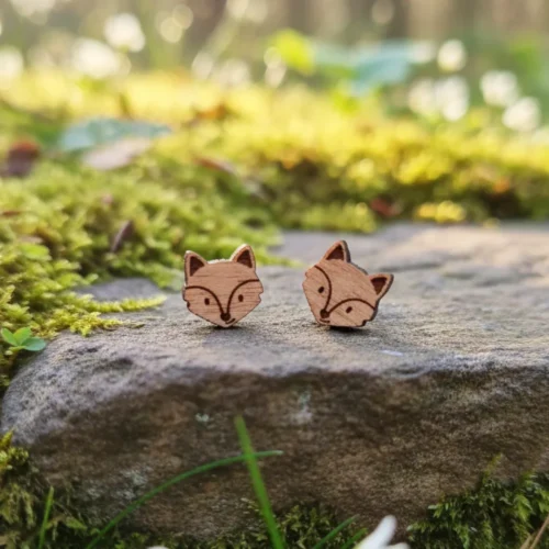 Clous-oreilles-bois-rox-renard Deux boucles d'oreilles têtes de renard en bois gravées posées sur pierre couverte de mousse verte lumineuse en forêt avec feuillage flou en arrière-plan
