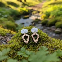 Boucles d'oreilles Peaks avec pendentifs blanc au relief de montagnes imprimées en 3D dans du plastique recyclé et pendentifs en bois marron pointus, sur mousse verte humeuse avec ruisseau de montagne visible en arrière-plan