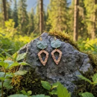 Boucles d'oreilles Peaks avec pendentifs gris clair vert au relief de montagnes imprimées en 3D dans du plastique recyclé et pendentifs en bois suisse pointus, sur roche grise avec petites plante vertes et arbres en arrière-plan